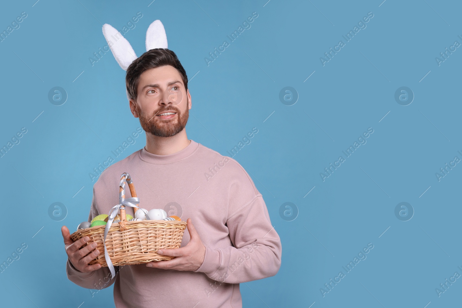 Photo of Happy man in cute bunny ears headband holding wicker basket with Easter eggs on light blue background. Space for text