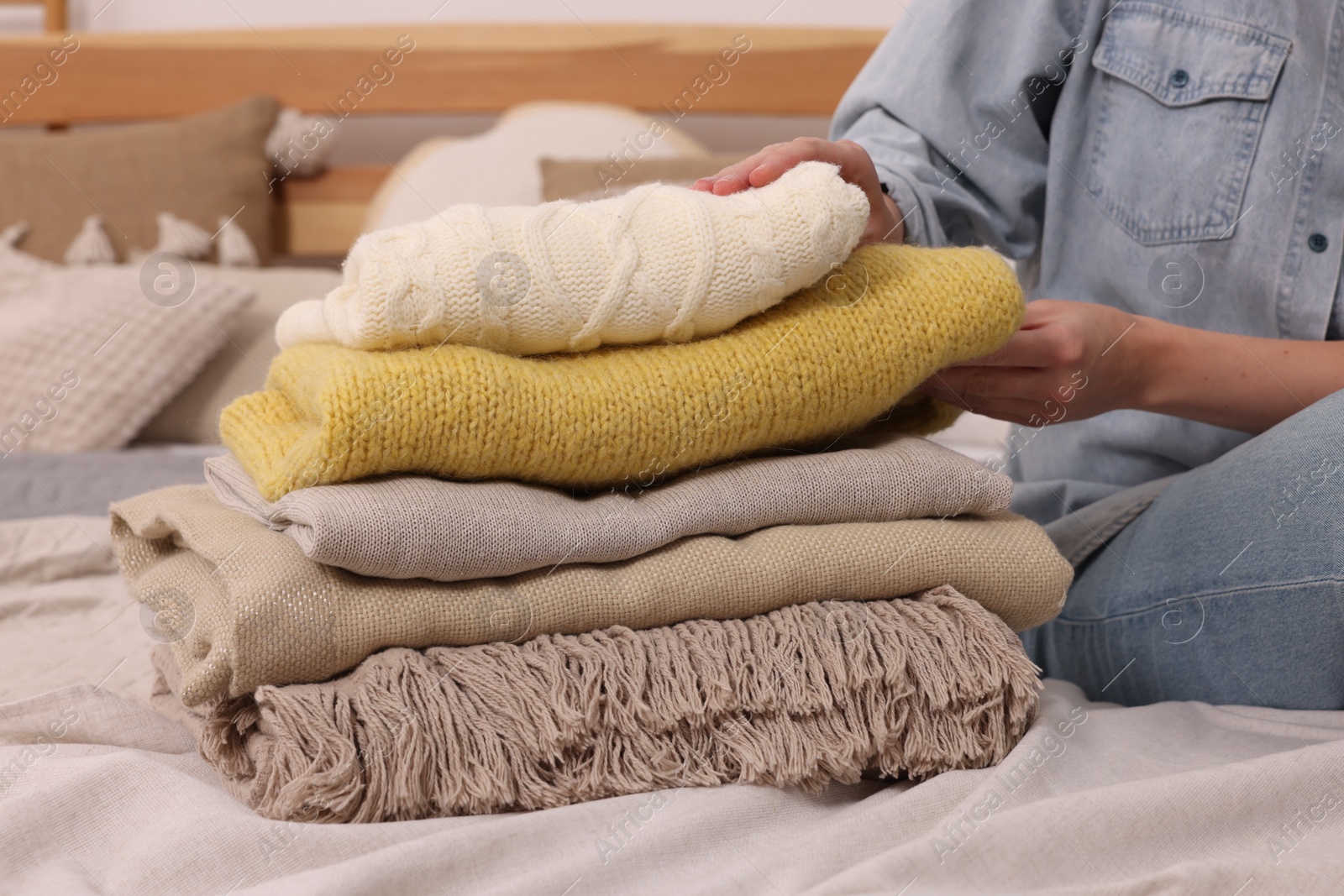 Photo of Woman with stack of different folded blankets and clothes on bed in room, closeup. Home textile