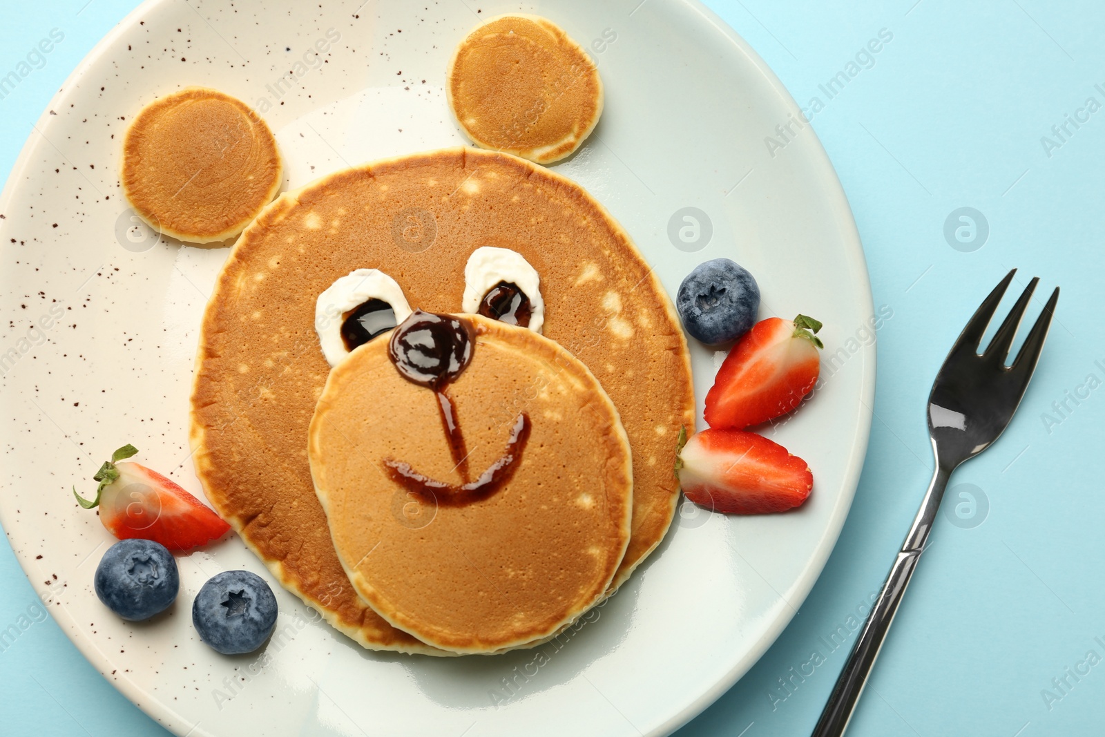 Photo of Creative serving for kids. Plate with cute bear made of pancakes and berries on light blue table, flat lay