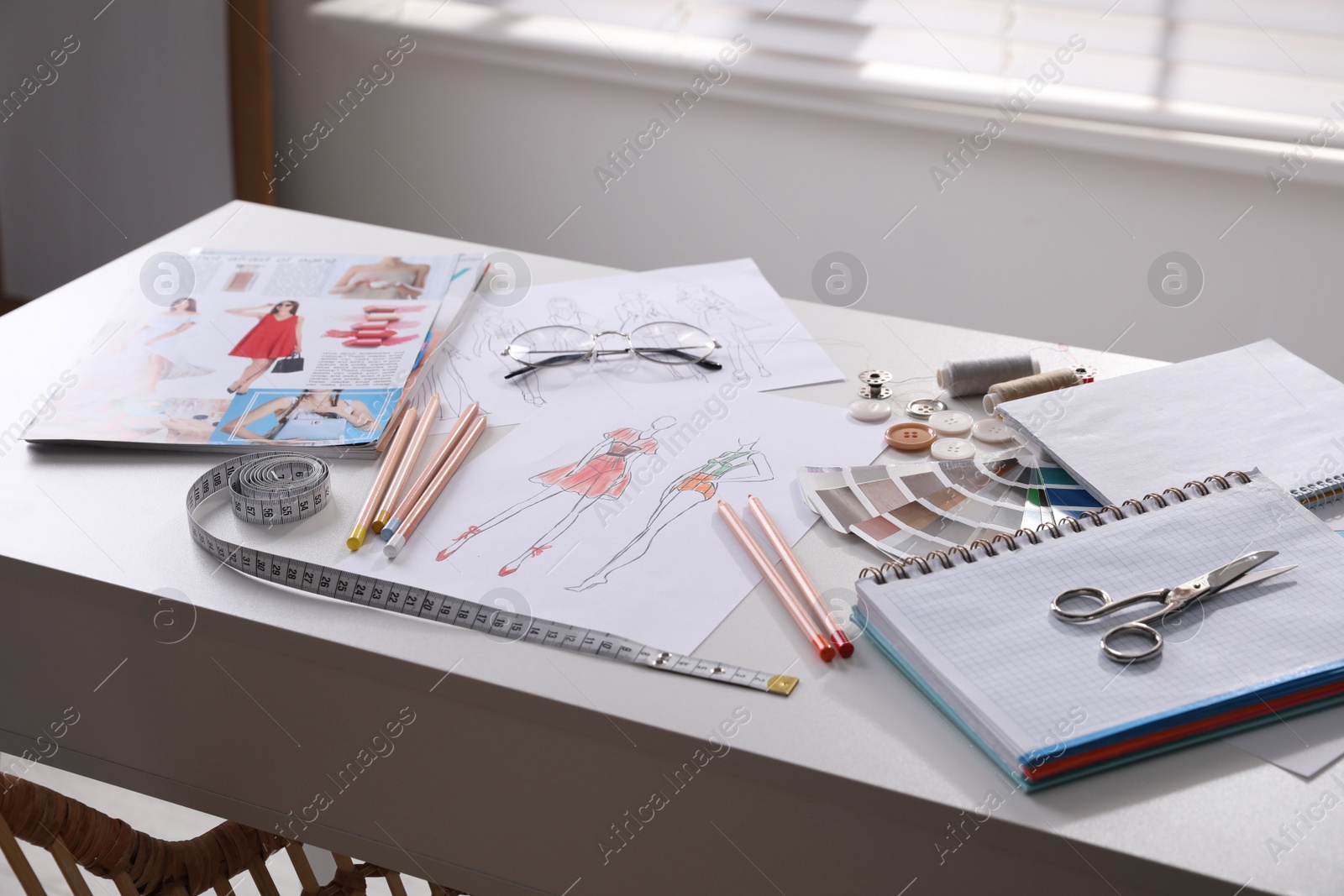 Photo of Sketches of clothes and different stuff on white table indoors. Fashion designer's workplace