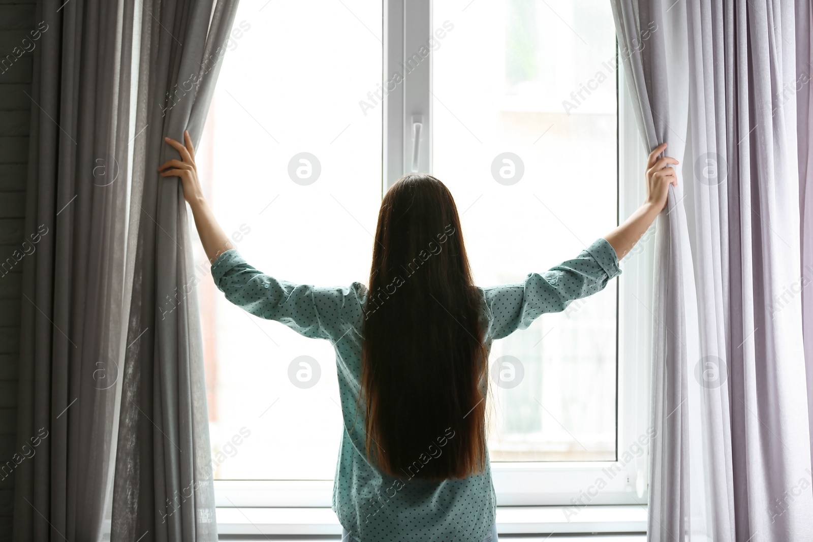 Photo of Young woman opening curtains in room near big beautiful window