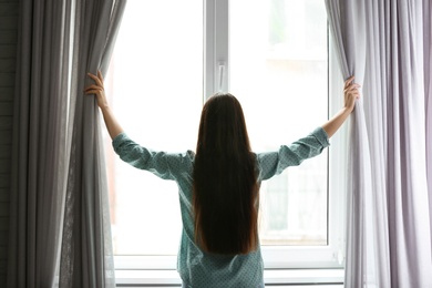 Young woman opening curtains in room near big beautiful window