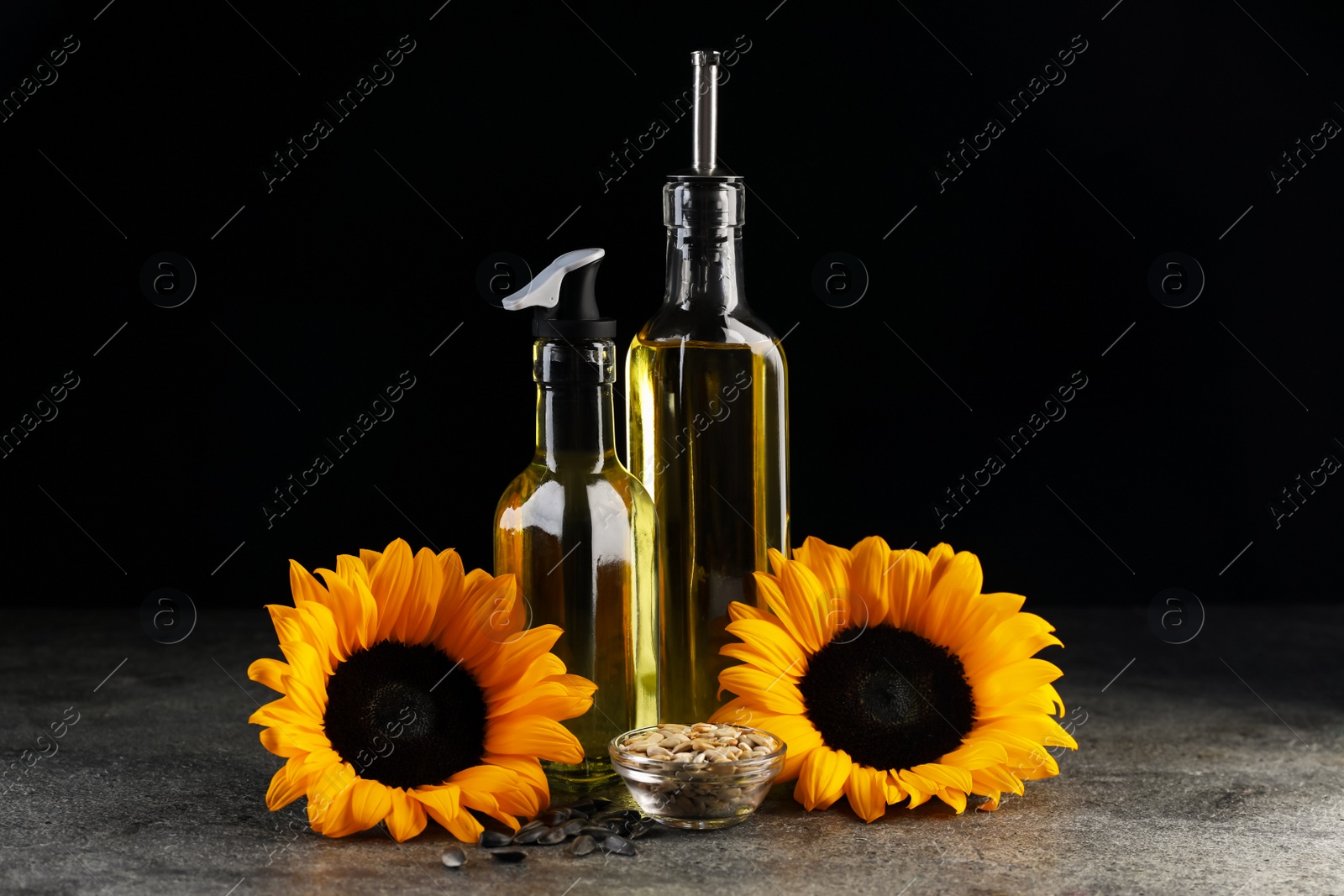 Photo of Sunflower cooking oil, seeds and beautiful flowers on grey table against black background