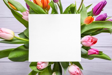 Photo of Beautiful bouquet of tulips with blank card on white wooden background, top view. Birthday celebration