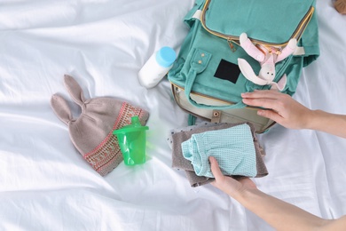 Photo of Woman packing baby accessories into maternity backpack on bed, top view