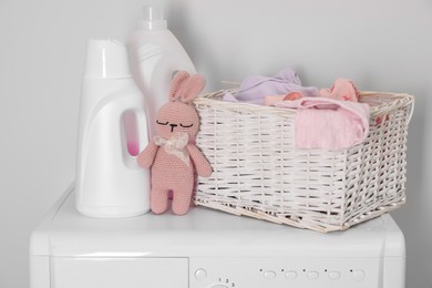 Photo of Baby clothes in wicker basket, laundry detergents and toy bunny on washing machine near light wall