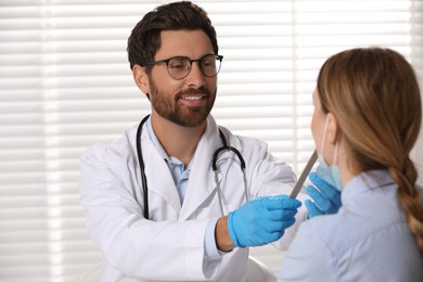 Photo of Smiling doctor examining woman`s oral cavity with tongue depressor indoors