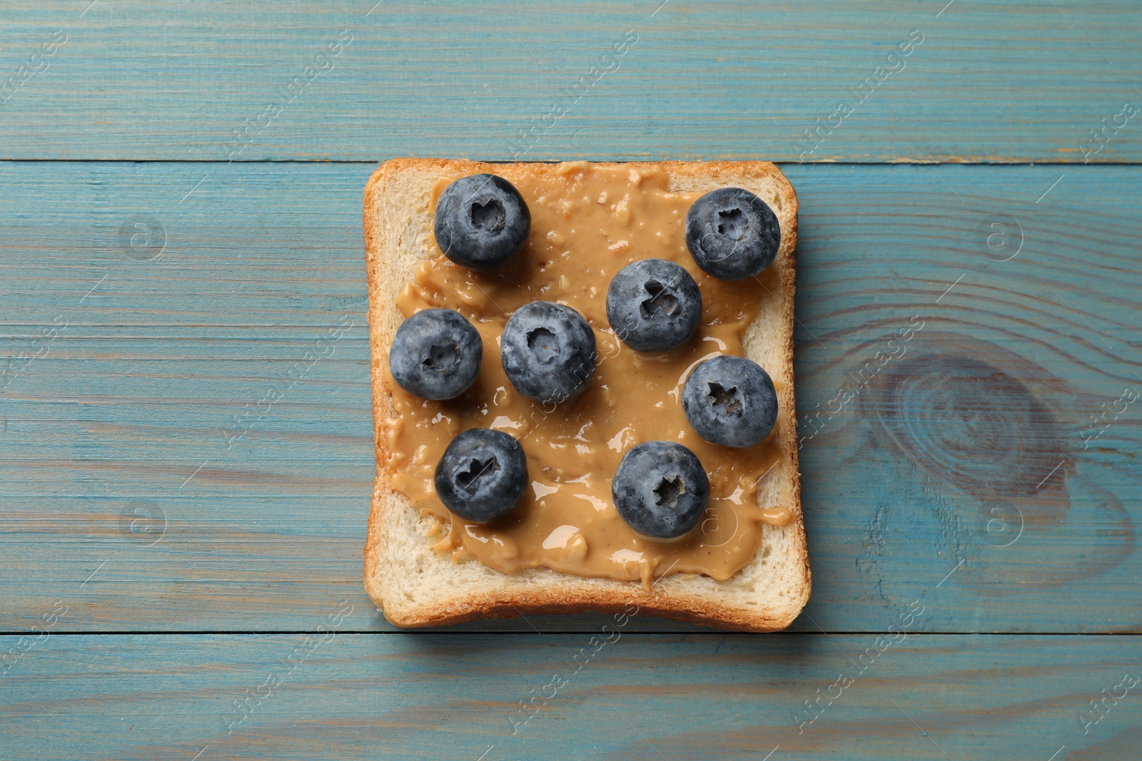 Photo of Delicious toast with peanut butter and blueberries on light blue wooden table, top view