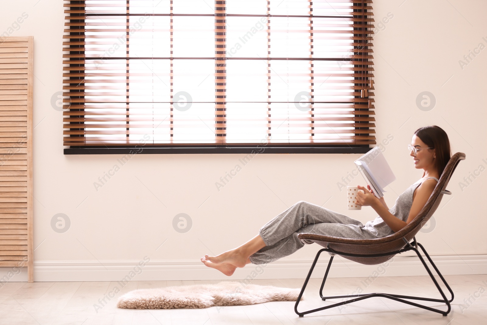 Photo of Young woman reading book near window with blinds at home. Space for text