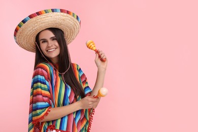 Photo of Young woman in Mexican sombrero hat and poncho dancing with maracas on pink background. Space for text