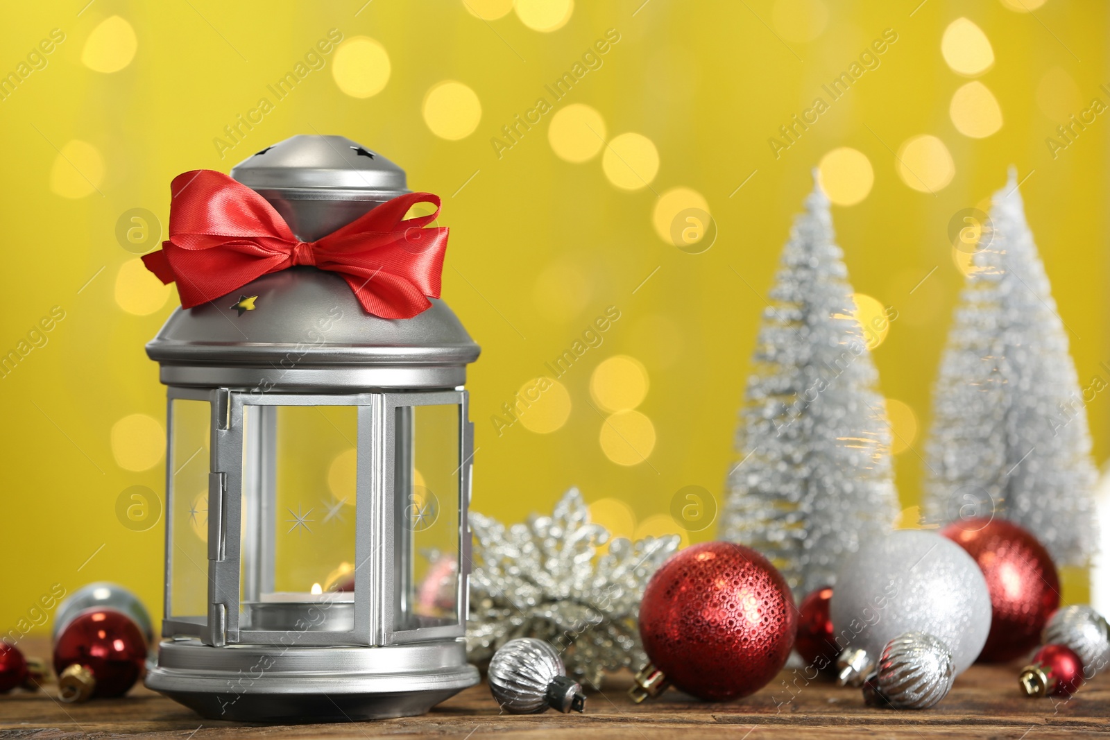 Photo of Christmas lantern with burning candle and festive ornaments on wooden table against blurred lights. Space for text