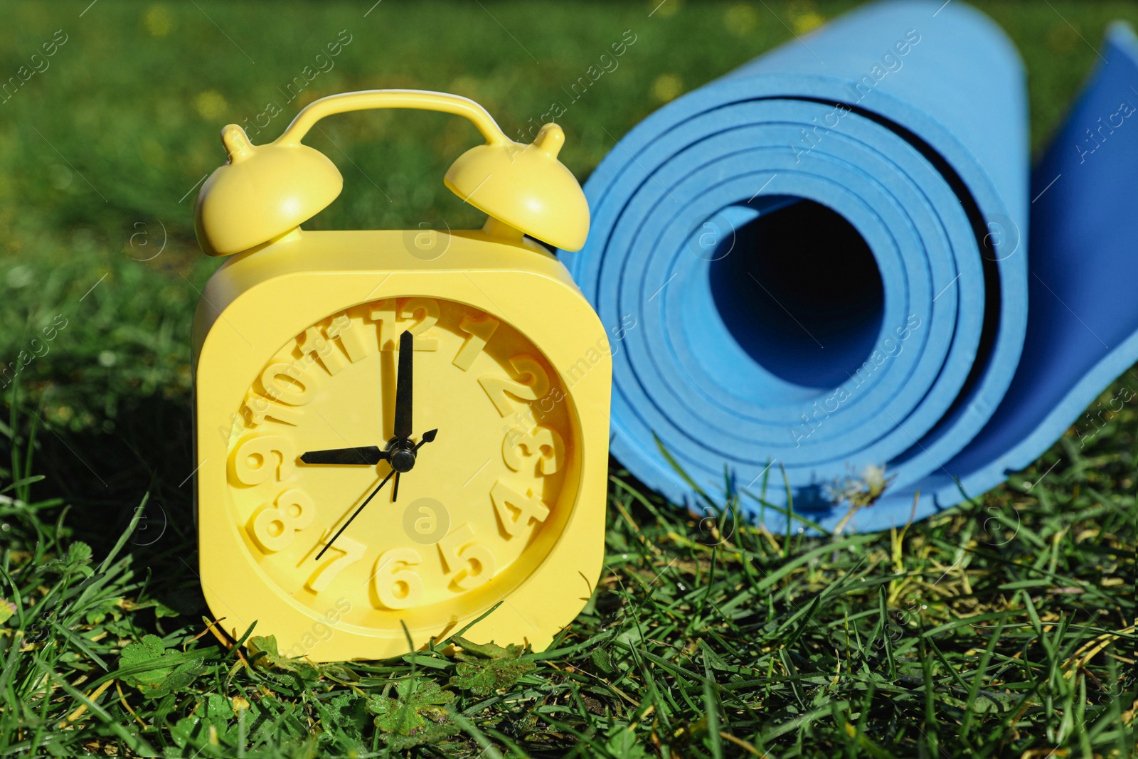 Photo of Alarm clock and fitness mat on green grass outdoors, closeup. Morning exercise