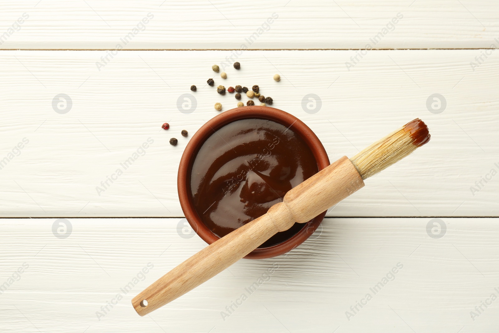 Photo of Tasty barbeque sauce in bowl, brush and peppercorns on white wooden table, top view