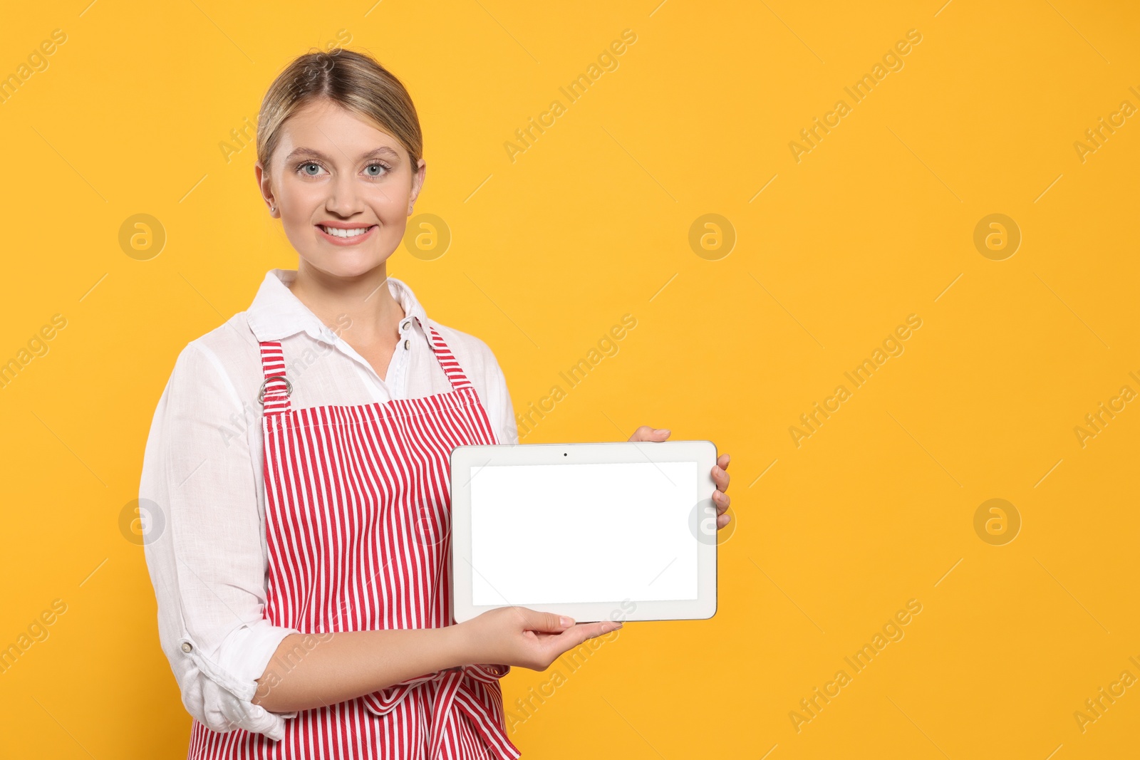 Photo of Beautiful young woman in clean striped apron with tablet on orange background. Space for text