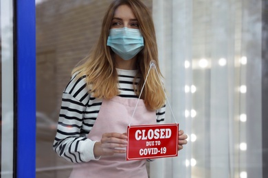 Photo of Business owner in mask hanging red sign with text Closed Due To Covid-19 onto glass door. Coronavirus quarantine