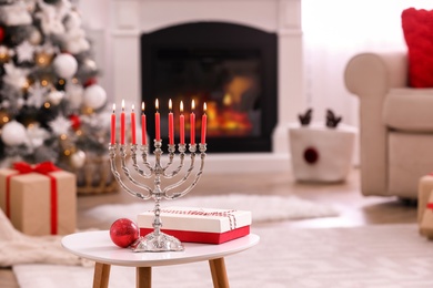 Silver menorah near Christmas ball and gift box on white table in room with fireplace, space for text. Hanukkah symbol