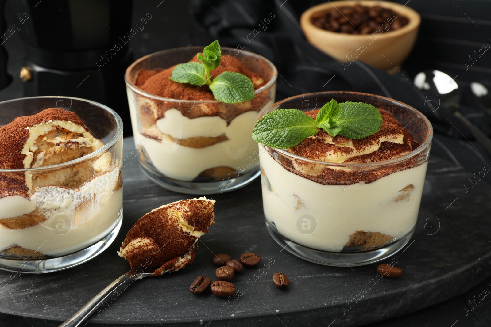 Photo of Delicious tiramisu in glasses, spoon, coffee beans and mint leaves on table