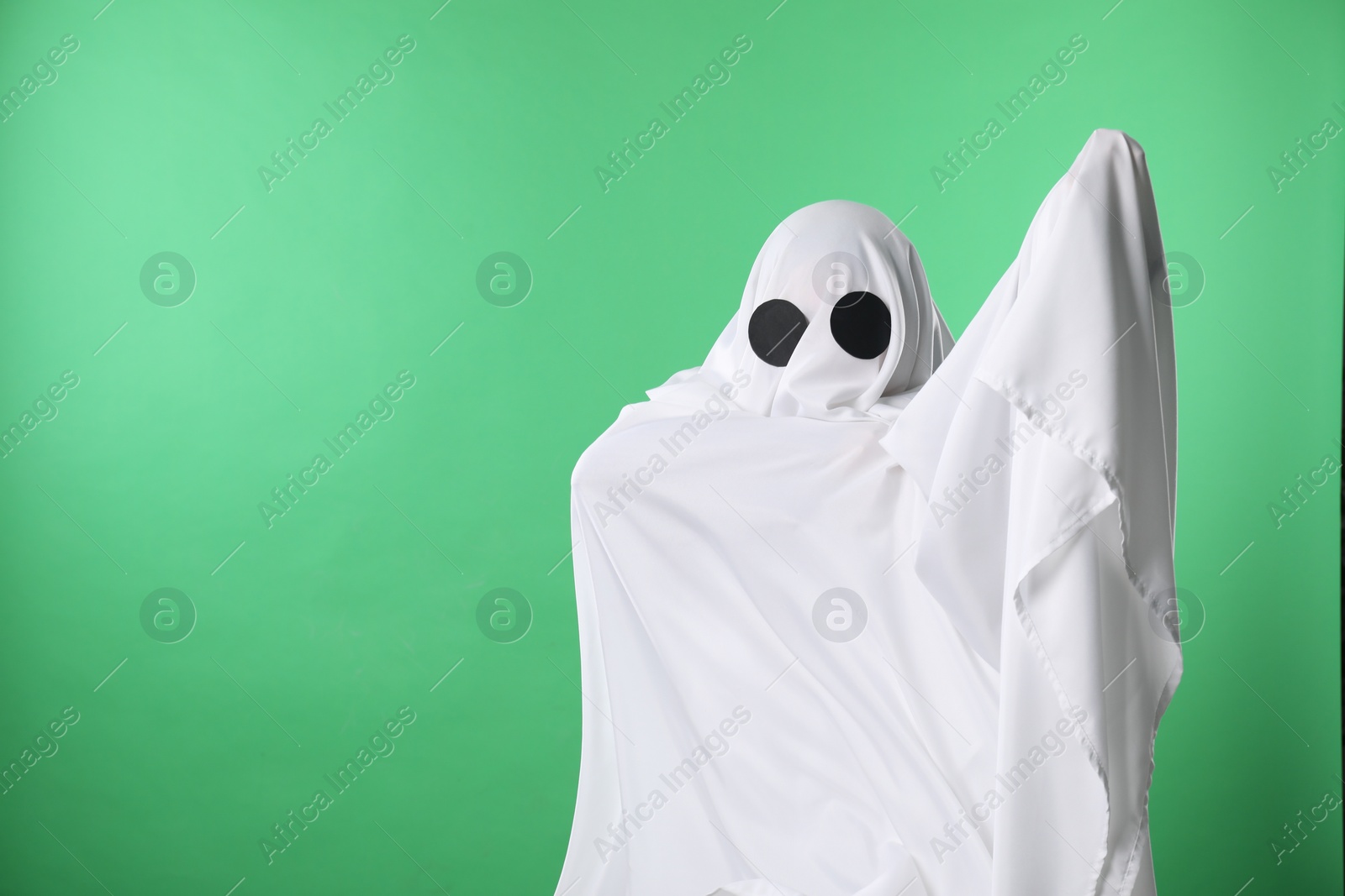 Photo of Creepy ghost. Person covered with white sheet on green background, space for text