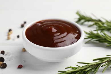 Photo of Tasty barbeque sauce in bowl, rosemary and spices on white wooden table, closeup