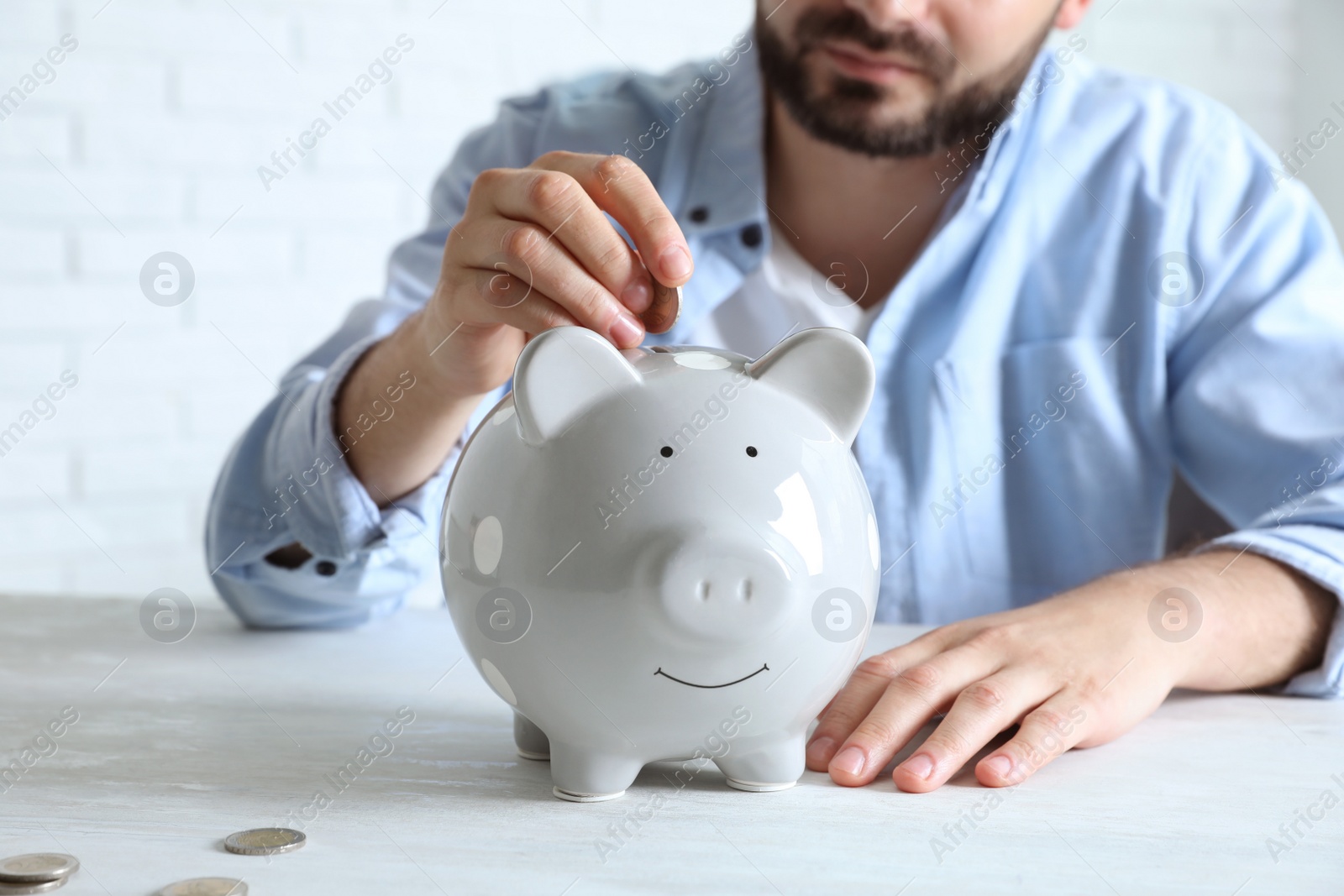 Photo of Man putting coin into piggy bank at white table indoors, closeup