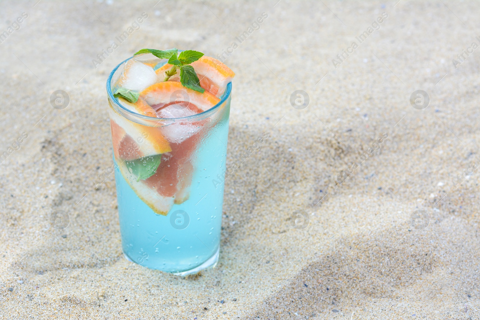 Photo of Glass of refreshing drink with grapefruit and mint on sandy beach, space for text