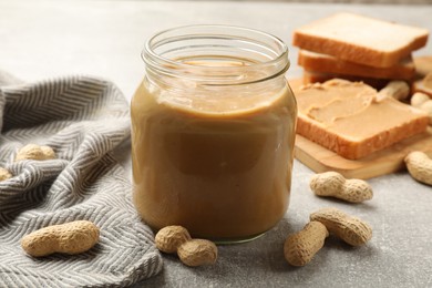 Photo of Tasty peanut butter in glass jar and peanuts on gray table, closeup