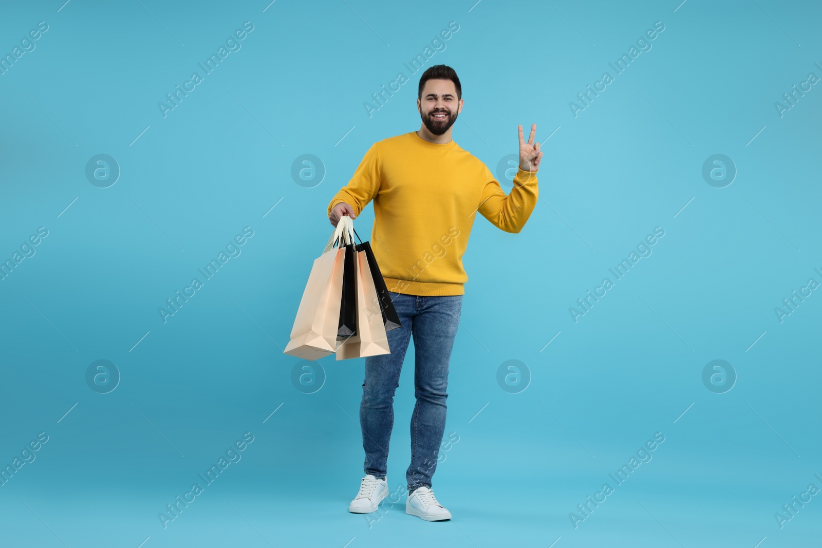 Photo of Happy man with many paper shopping bags showing peace sign on light blue background