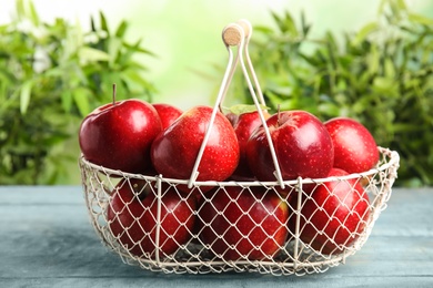 Photo of Metal basket with ripe juicy red apples on blue wooden table against blurred background