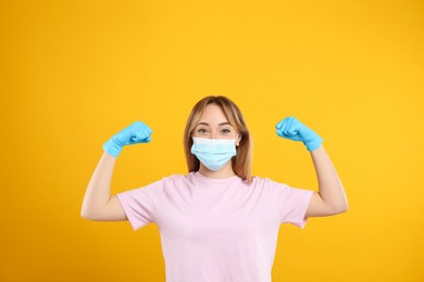 Photo of Woman with protective mask and gloves showing muscles on yellow background. Strong immunity concept