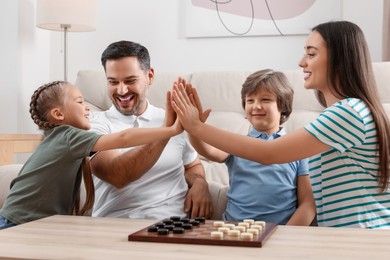 Photo of Playing checkers. Parents giving high five to children at table in room