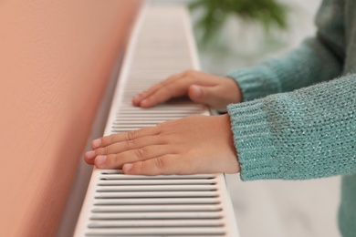 Photo of Child warming hands on heating radiator indoors, closeup