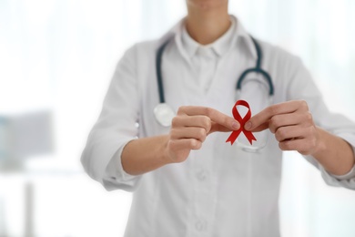 Photo of Doctor holding red ribbon on blurred background, closeup. AIDS awareness month