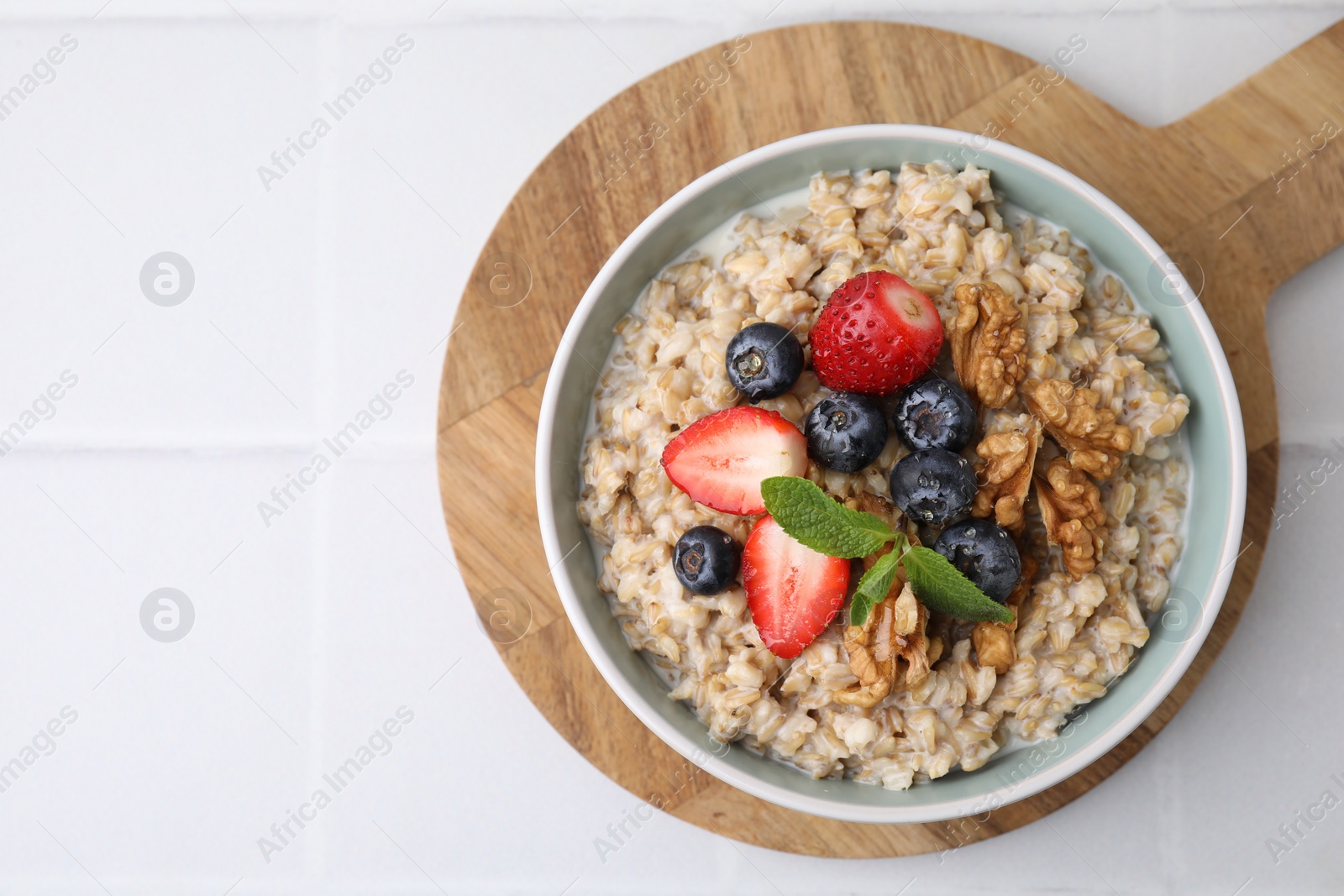 Photo of Tasty oatmeal with strawberries, blueberries and walnuts in bowl on white tiled table, top view. Space for text