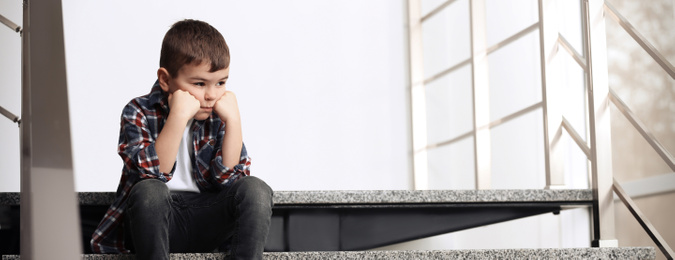 Sad little boy sitting on stairs indoors, banner design with space for text. Time to visit child psychologist