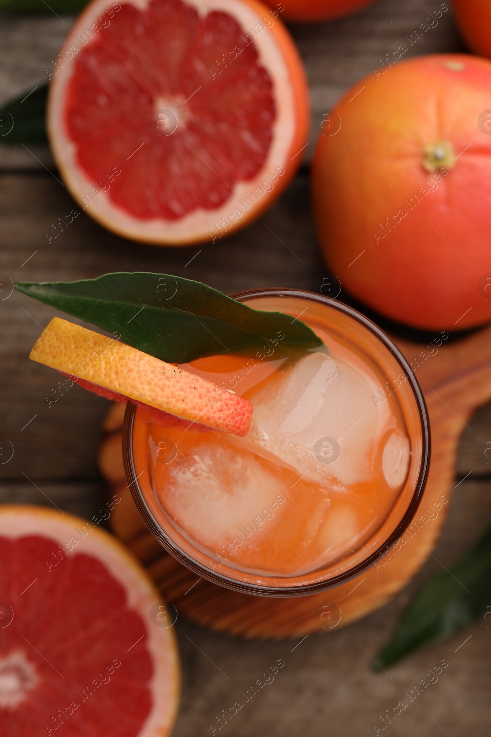 Photo of Tasty grapefruit drink with ice and leaf in glass surrounded by fresh fruits on wooden table, flat lay