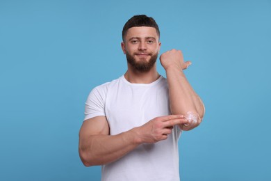 Handsome man applying body cream onto his elbow on light blue background