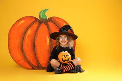 Photo of Cute little girl with candy bucket and decorative pumpkin wearing Halloween costume on yellow background