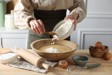 Photo of Making dough. Woman adding flour into bowl at wooden table in kitchen, closeup