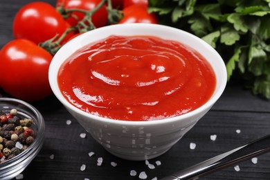 Photo of Delicious ketchup in bowl and spices on black wooden table, closeup. Tomato sauce
