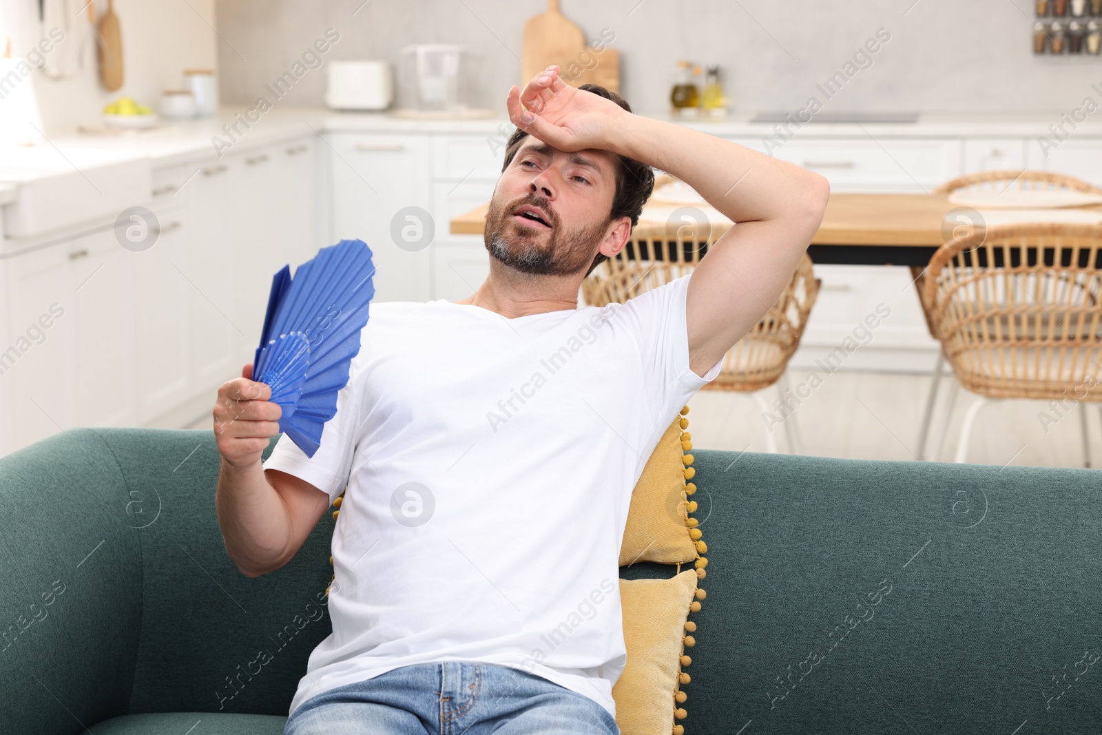 Photo of Bearded man waving blue hand fan to cool himself on sofa at home