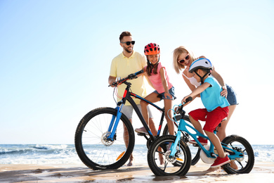 Happy parents teaching children to ride bicycles on sandy beach near sea