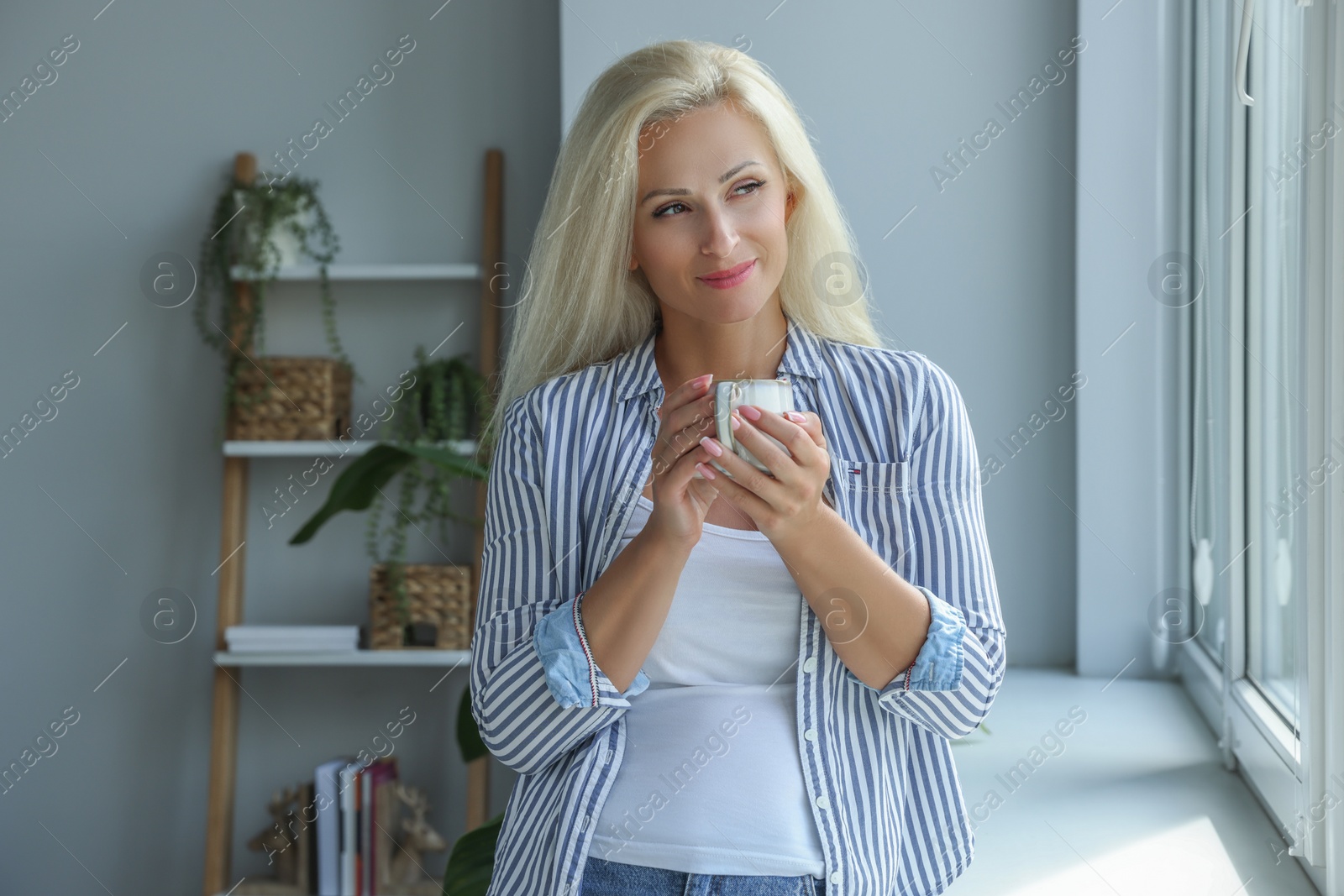 Photo of Beautiful woman with cup of hot drink near window at home