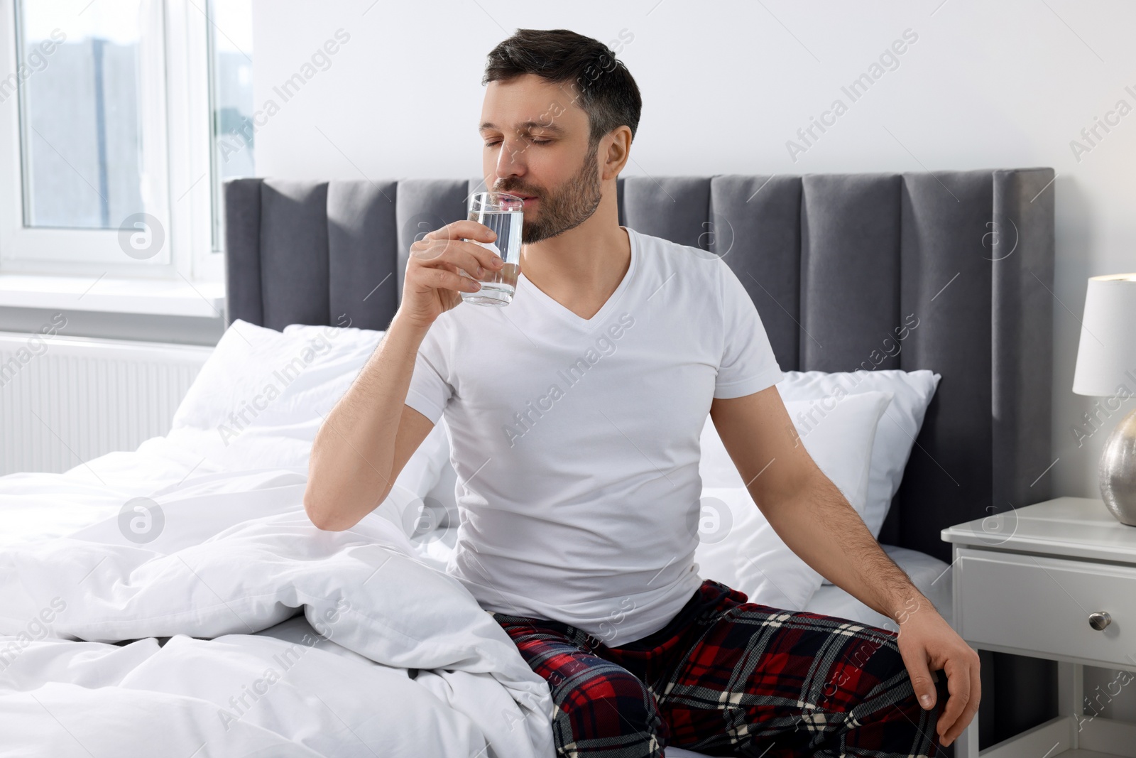 Photo of Handsome man drinking water from glass on soft bed at home in morning
