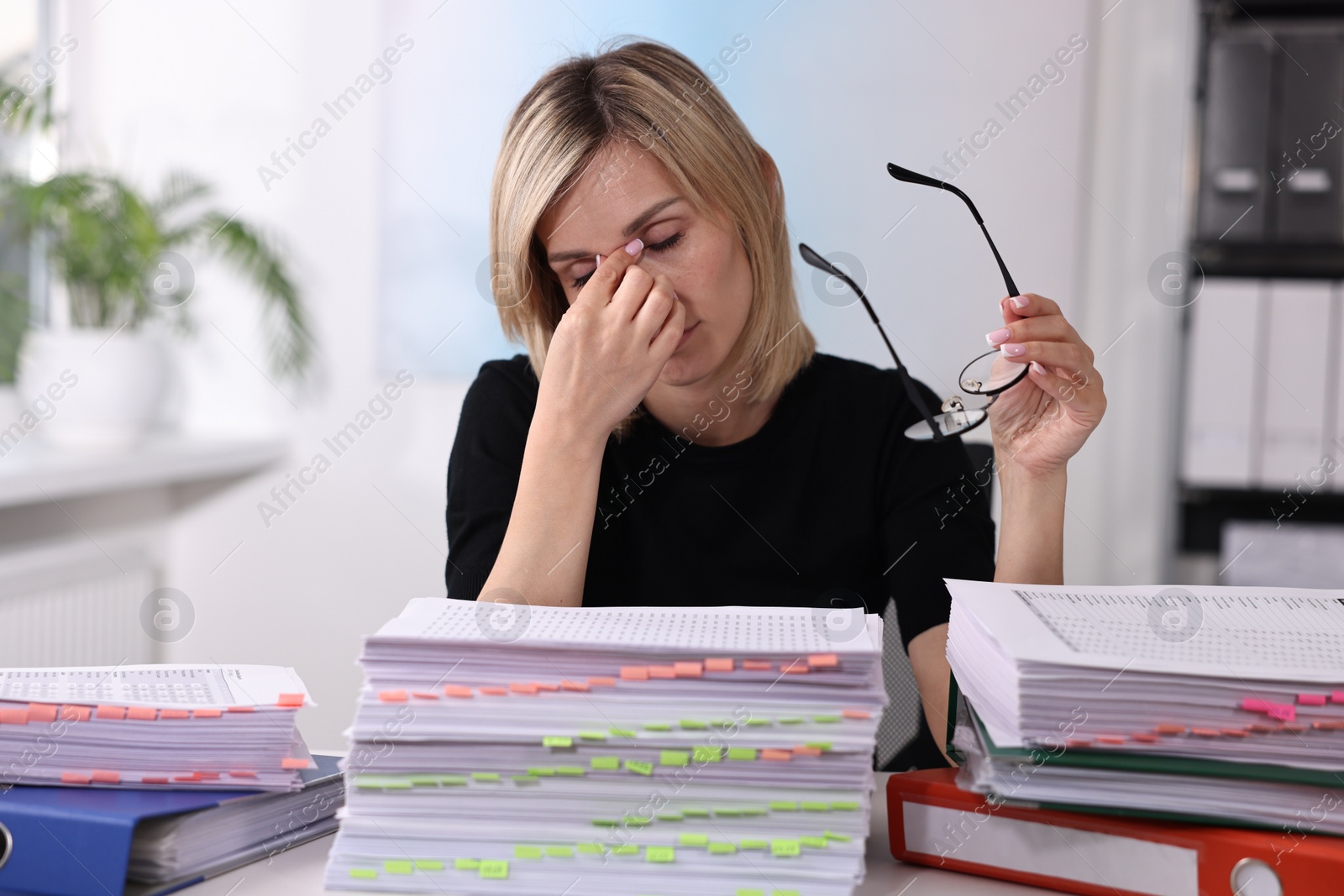 Photo of Overwhelmed woman sitting at table with stacks of documents and folders in office