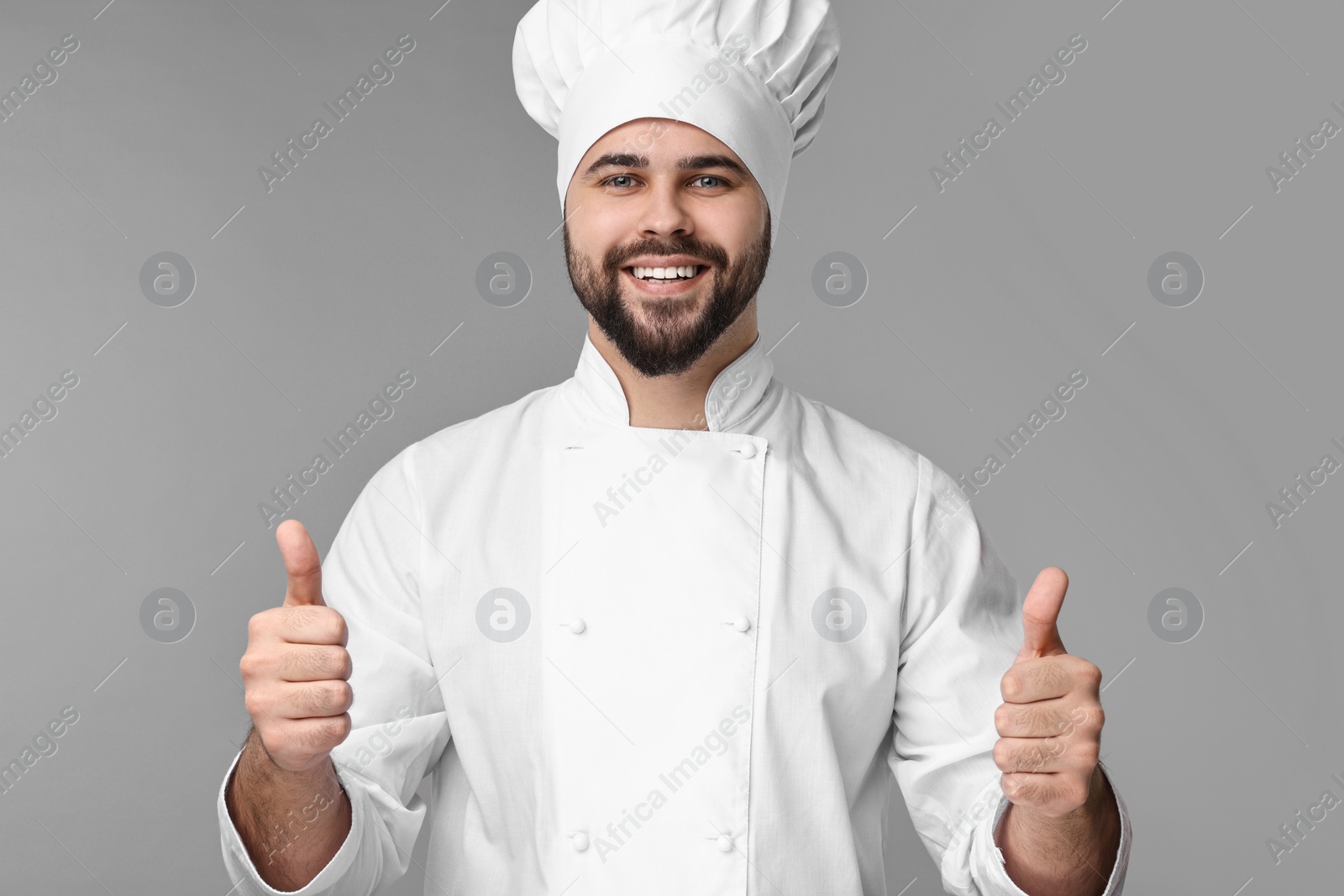 Photo of Happy young chef in uniform showing thumbs up on grey background