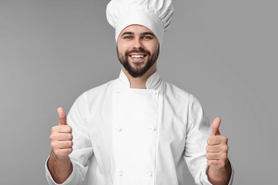 Happy young chef in uniform showing thumbs up on grey background