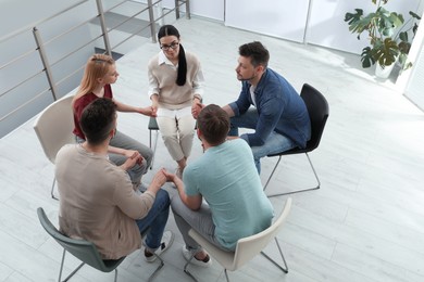 Photo of Psychotherapist and group of drug addicted people holding hands together at therapy session indoors, above view