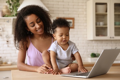 Photo of African-American woman and her baby with laptop in kitchen. Happiness of motherhood