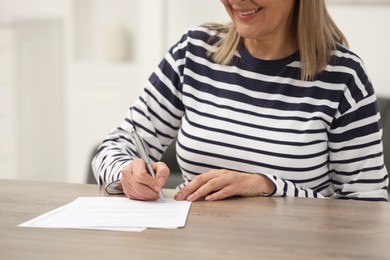 Photo of Smiling senior woman signing Last Will and Testament at wooden table indoors, closeup