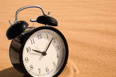 Photo of Black alarm clock on sand in desert, closeup. Space for text